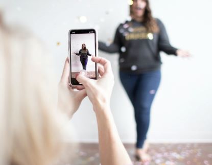 Woman stands near a wall in casual jeans with glitter floating down around her. In the forefront, another women holds up a cell phone and is shooting video. 